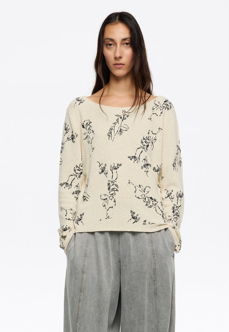 Hellbeige strukturierte Strickjacke mit schwarzem Blumenmuster, weit ausgeschnittenem Kragen und langenÄrmeln, kombiniert mit grauen, locker sitzenden Hosen.