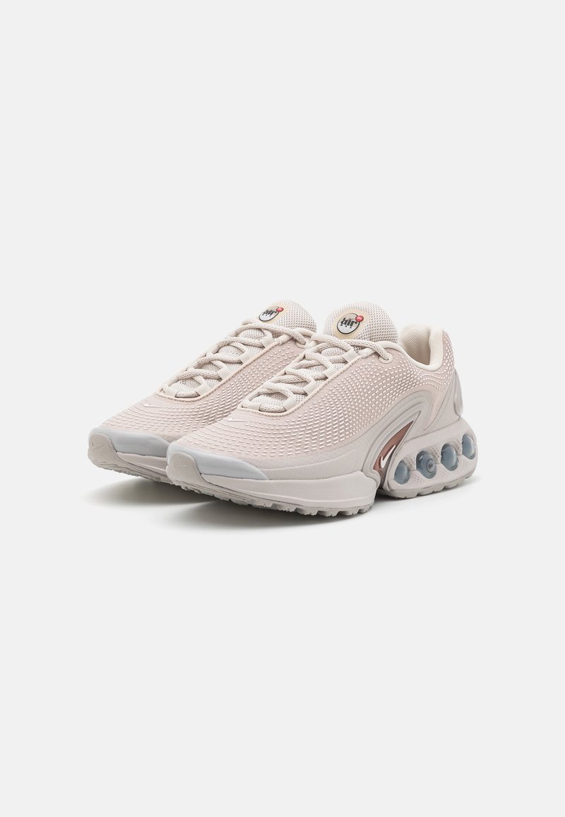 Paire de baskets de sport beige clair en tissu texturé, avec lacets et coussinets d'amorti transparents sur la semelle, posées sur un fond blanc.