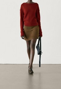 Pull rouge tricoté avec manches larges, associé à une mini-jupe en daim marron. Sac à bandoulière noir, chaussures à motifs, marchant sur une surface neutre.