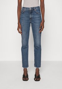 Siniset farkut, joissa suora lahje, korkea vyötärö ja rispaantunut helma. Klassinen viiden taskun design ja kontrastitikkaukset.