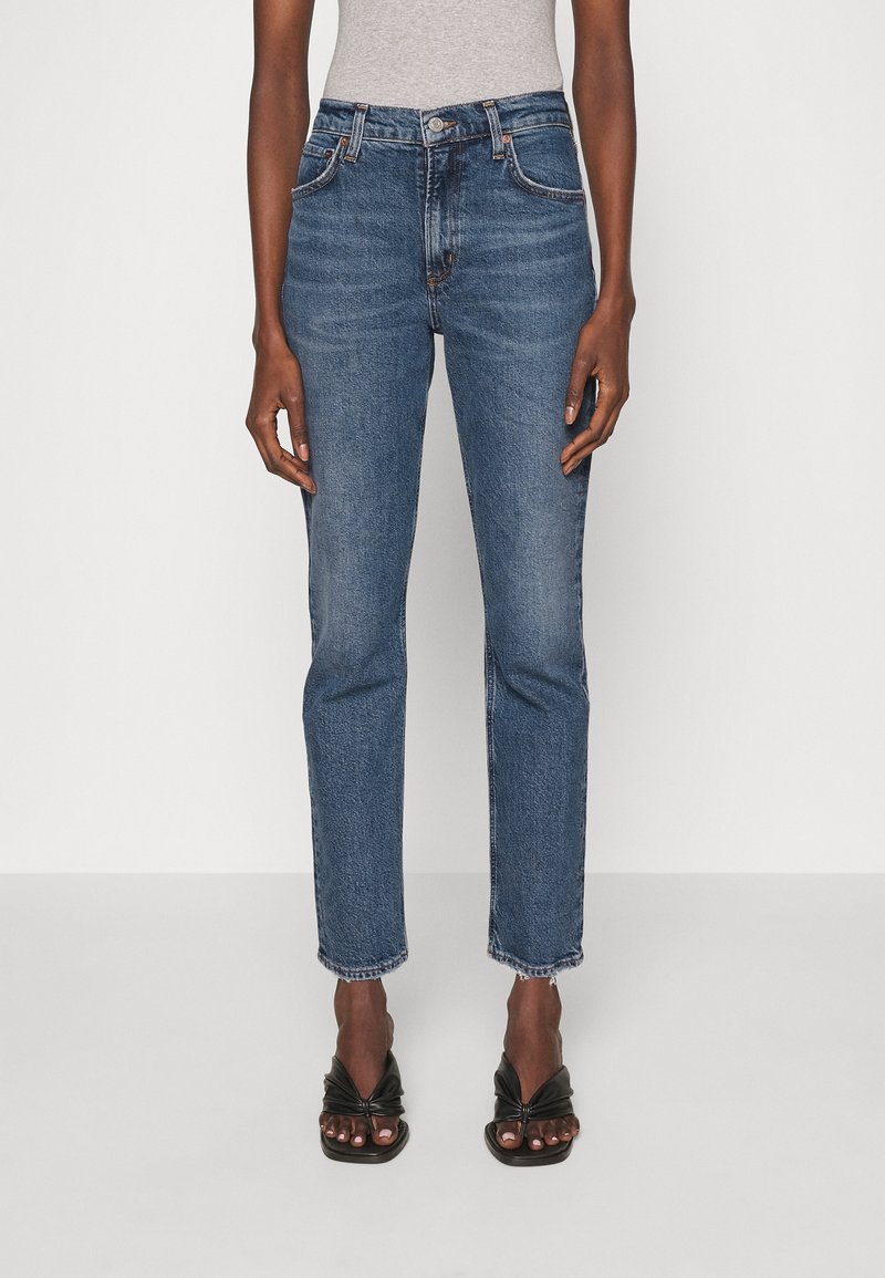 Siniset farkut, joissa suora lahje, korkea vyötärö ja rispaantunut helma. Klassinen viiden taskun design ja kontrastitikkaukset.