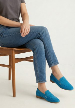 Person trägt blaue Wildleder-Loafers und blaue Jeans, sitzt mit überkreuzten Beinen auf einem Holzstuhl vor einem einfarbigen Hintergrund.
