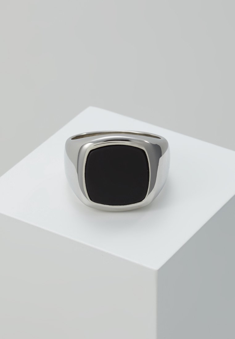 Bague chevalière en argent avec une pierre onyx noire lisse et carrée au centre, exposée sur un piédestal géométrique blanc.