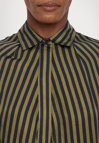 Gros plan d’une personne portant une chemise à col boutonné avec des rayures verticales noires et vert olive.