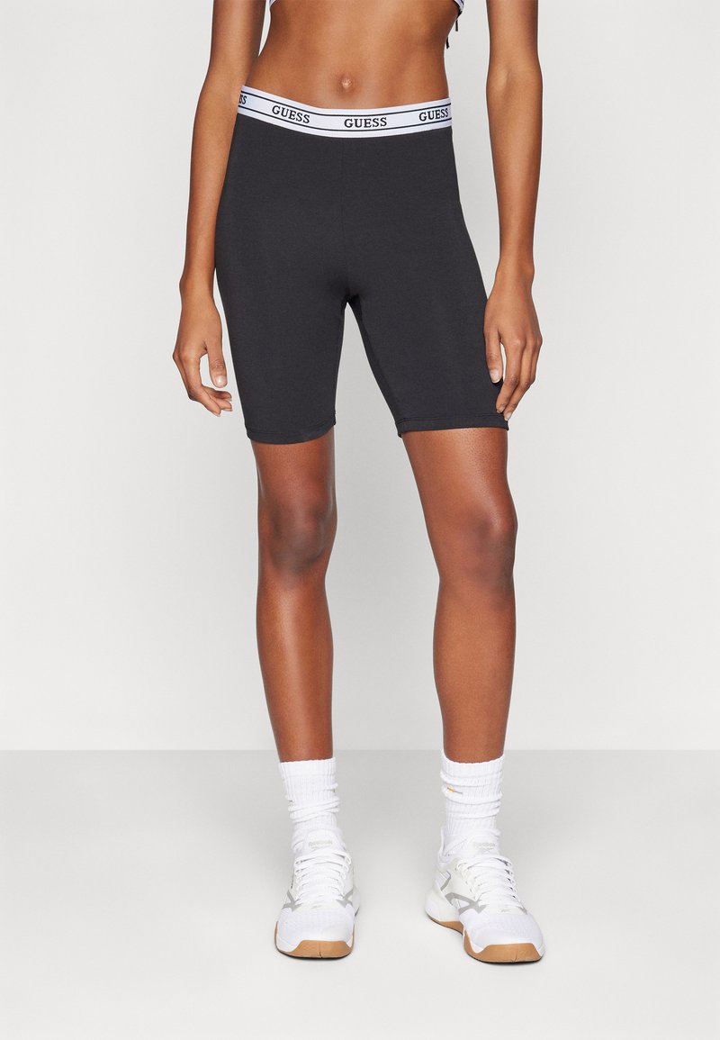 Shorts de vélo en coton noir avec une ceinture logo "GUESS" blanche, présentant un design ajusté et une longueur jusqu'au mi-thigh.