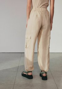 Lys beige cargo-bukser med elastisk talje, sidelommer og løs pasform, parret med sorte platåsandaler med grønne detaljer.