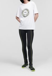 Witte t-shirt met zwart rond logo en groene accenten, gecombineerd met zwarte leggings met neon groene zijkanten en zwarte sportschoenen.