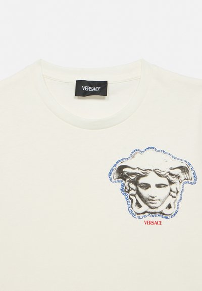 Λευκό μπλουζάκι με στρογγυλή λαιμόκοψη, με ετικέτα Versace και έμβλημα κεφαλής Μέδουσας σε μαύρο και άσπρο, περιγραμμένο με μπλε χρώμα, πάνω από κόκκινο κείμενο Versace.