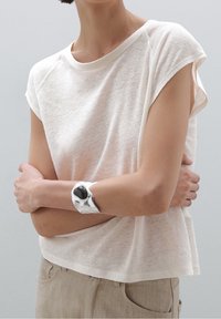 Lys beige, kortærmet t-shirt lavet af et tekstureret stof, parret med et sølvarmbånd med en sort stenaccent.