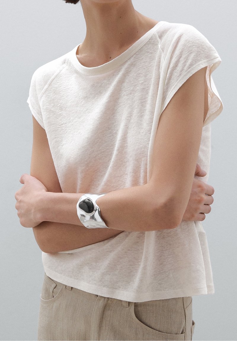 Lys beige, kortærmet t-shirt lavet af et tekstureret stof, parret med et sølvarmbånd med en sort stenaccent.