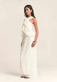 Robe en lin blanc avec un ourlet asymétrique à volants, design sans manches, tissu superposé et silhouette fluide, complétée par un bracelet en or.