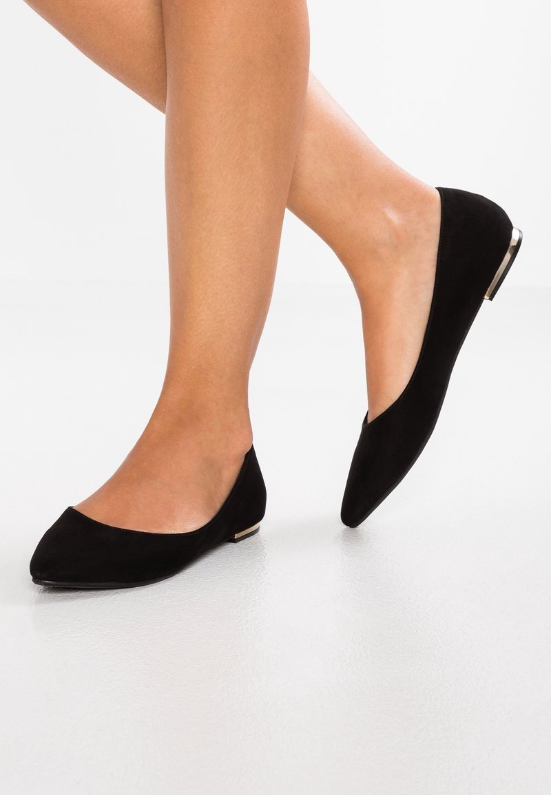 Femme portant des ballerines en daim noir à bout pointu avec un détail métallique sur le talon, debout sur un sol blanc texturé.
