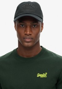 Man wearing a black Superdry cap and a dark green Superdry t-shirt with yellow logo lettering on the chest, facing forward.
