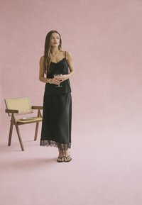 Woman in a black dress holding a cocktail glass, standing next to a wooden chair with a tray of glasses on a pink floor and wall.
