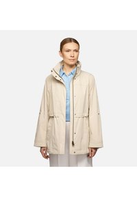 Giacca beige leggera con colletto alto, bottoni a scatto, vita con cordino e due tasche frontali. Indossata sopra una camicia azzurro chiaro.