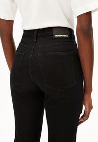Jean noir taille haute en denim avec une texture lisse, dotés de poches arrière plaquées et d'une étiquette ARMADANGELS sur la taille.