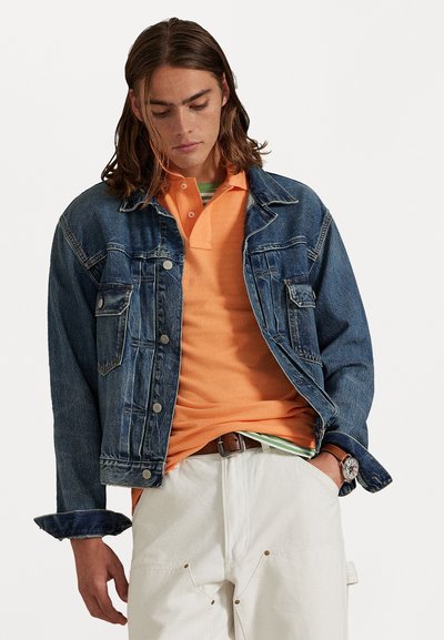 Νέος άνδρας με μακριά μαλλιά, φορώντας τζιν μπουφάν πάνω από πορτοκαλί πόλο και λευκά παντελόνια, στέκεται με το ένα χέρι στην τσέπη και κοιτάζει κάτω.