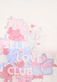 Ανοιχτόχρωμο T-shirt με φλοράλ σχέδιο σε ροζ και μπλε αποχρώσεις, με το κείμενο "SELF LOVE CLUB" σε έντονα λευκά γράμματα. Διακοσμημένο με στρας.
