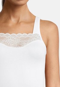 Top bianco senza maniche realizzato in morbido tessuto, con un bordo decorativo in pizzo lungo il colletto. Le spalline sono sottili e il design è a girocollo.
