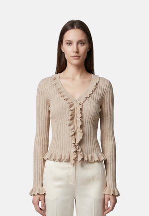 Femme portant un cardigan côtelé beige avec des volants le long de la ligne de boutons, des poignets et de l'ourlet, associé à un pantalon blanc taille haute, debout devant un fond blanc.