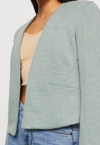 Blazer vert clair court en tissu texturé, avec un col cranté et une ouverture sur le devant. Porté sur un haut beige et un jean bleu.