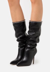 Botas negras de cuero hasta la rodilla con diseño holgado, punta puntiaguda y un puño decorativo con acentos de brillantes. Tacón de aguja.