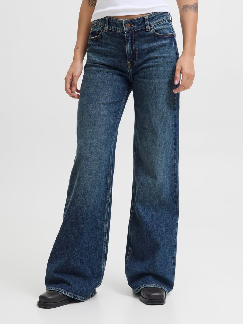 Asmuo dėvi tamsiai mėlynas plačias džinsų kelnes ir juodus batus, stovi priešais vientisą baltą fono sieną, matomi rankų tatuiruotės.