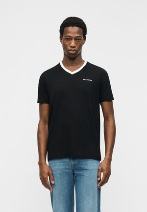 Jeune homme portant un T-shirt noir à col en V avec un liseré blanc et un petit logo blanc sur la poitrine, assorti à un jean bleu clair, debout devant un fond blanc.