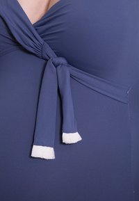 Marineblauwe zwemoutfit met een wikkeldesign, een voordelige knoop en witte franjes aan de uiteinden. Glad textuur, elastisch materiaal.