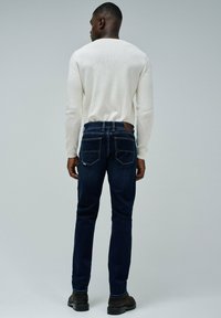 Jeans de mezclilla azul oscuro con un corte estructurado, que cuentan con bolsillos traseros con detalles de costura y una etiqueta de cintura de cuero marrón.
