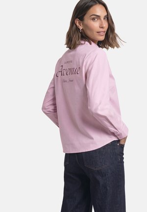 Kvinde med skulderlangt hår iført en lyserød skjorte med knapper med teksten "La Petite Avenue Paris, France" på ryggen og mørkeblå jeans.