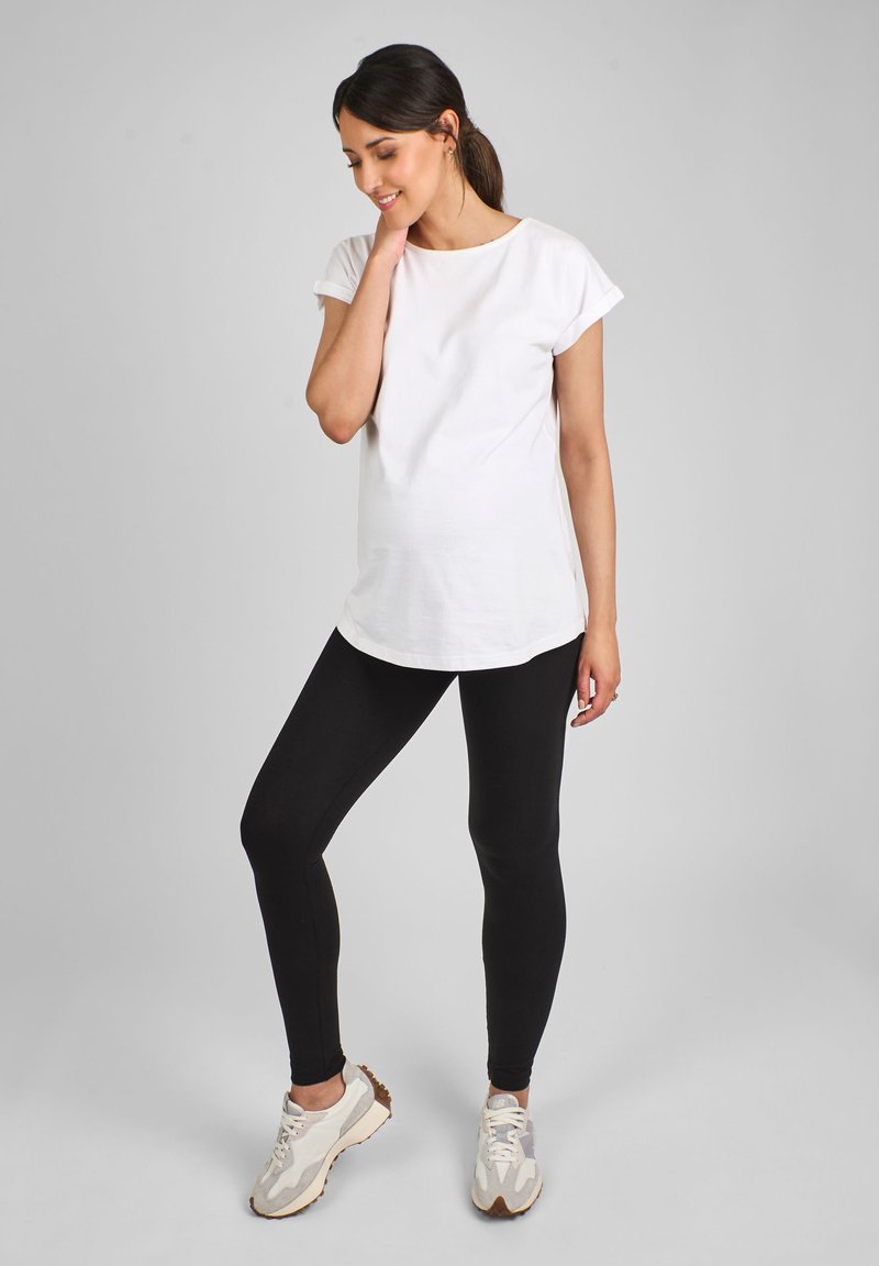 Kvinde med mørkt hår i hestehale iført en hvid t-shirt, sorte leggings og hvide sneakers, stående med en hånd på nakken.