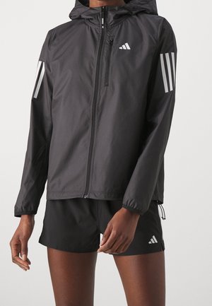 Homme portant une veste zippée à capuche noire avec des bandes réfléchissantes sur les manches et un logo sur la poitrine, associée à un short noir.