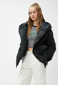 Ung kvinde med langt bølget hår iført en sort oversized pufferjakke, stribet cropped top og hvide cargo-bukser foran en ensfarvet baggrund.