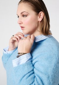 Lichtblauwe oversized trui met een geribbelde textuur, gedragen over een lichtblauw-wit gestreept overhemd met een kraag en metallic accenten.