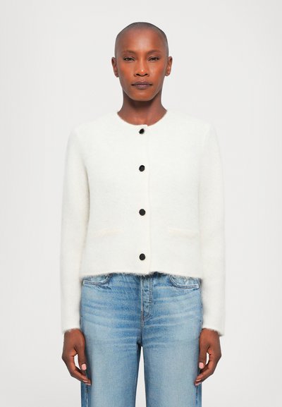 White, cropped cardigan with a round neckline, black buttons, and two front pockets. Soft texture and relaxed fit. Paired with blue jeans.