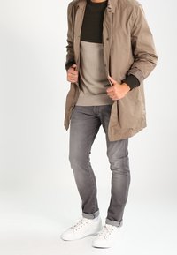 Beige lång jacka, olivgrön crewneck, grå jeans, vita sneakers. Jackan har knäppning, ribbade ärmmuddar och en avslappnad passform.