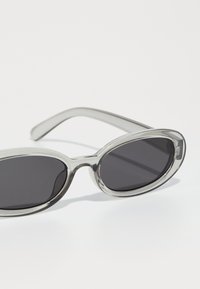 Gafas de sol con montura gris transparente, lentes ovalados con tinte oscuro y patillas delgadas, que exhiben un diseño minimalista y una textura suave.