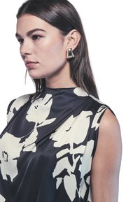 Blusa senza maniche in satin con motivo floreale bianco e nero. Il modello indossa un orecchino scultoreo dorato. Texture liscia, design elegante.