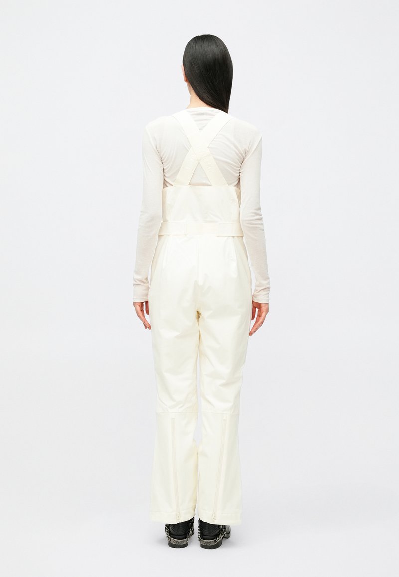 Tuta impermeabile color crema con bretelle regolabili incrociate sulla schiena, gambe dei pantaloni svasate e cerniere alle caviglie. Tessuto dalla texture liscia e design pratico.