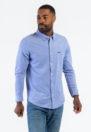 Chemise boutonnée bleu clair avec col, manches longues et ourlet arrondi. Présente un petit motif de logo au niveau de la poche poitrine.