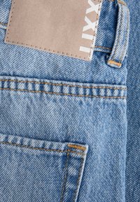 Ανοιχτό μπλε τζιν με πίσω τσέπη, πορτοκαλί ραφές, θηλιά για ζώνη και μερικώς ορατή μπεζ ετικέτα επωνυμίας.