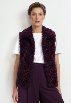 Donna con capelli corti che indossa una t-shirt bianca, un gilet testurizzato viola scuro e pantaloni a vita alta viola scuro abbinati su sfondo bianco.