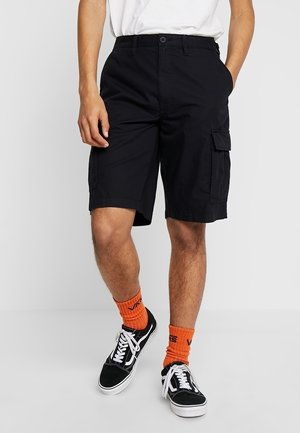 Mężczyzna ubrany w czarne szorty cargo, białą koszulę, jaskrawe pomarańczowe skarpetki Vans i czarne sneakersy Vans, stojący na tle jednolitym.