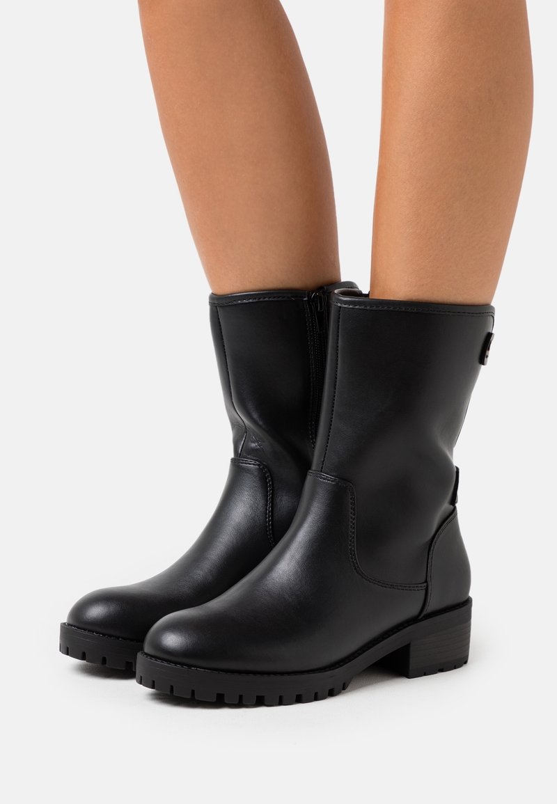 Bottes en cuir noir mi-mollet avec fermetures éclair latérales et talons bas épais, portées sur des jambes nues sur fond blanc.