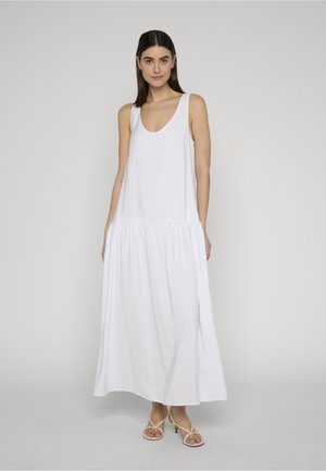 Donna con capelli lisci e scuri che indossa un vestito lungo bianco senza maniche e sandali beige con tacco a fascia, in piedi davanti a uno sfondo semplice.