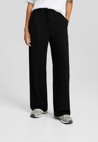 Svarta byxor med vida ben och hög midja, med veck fram; kombineras med grå sneakers med en sål av mesh och gummi.