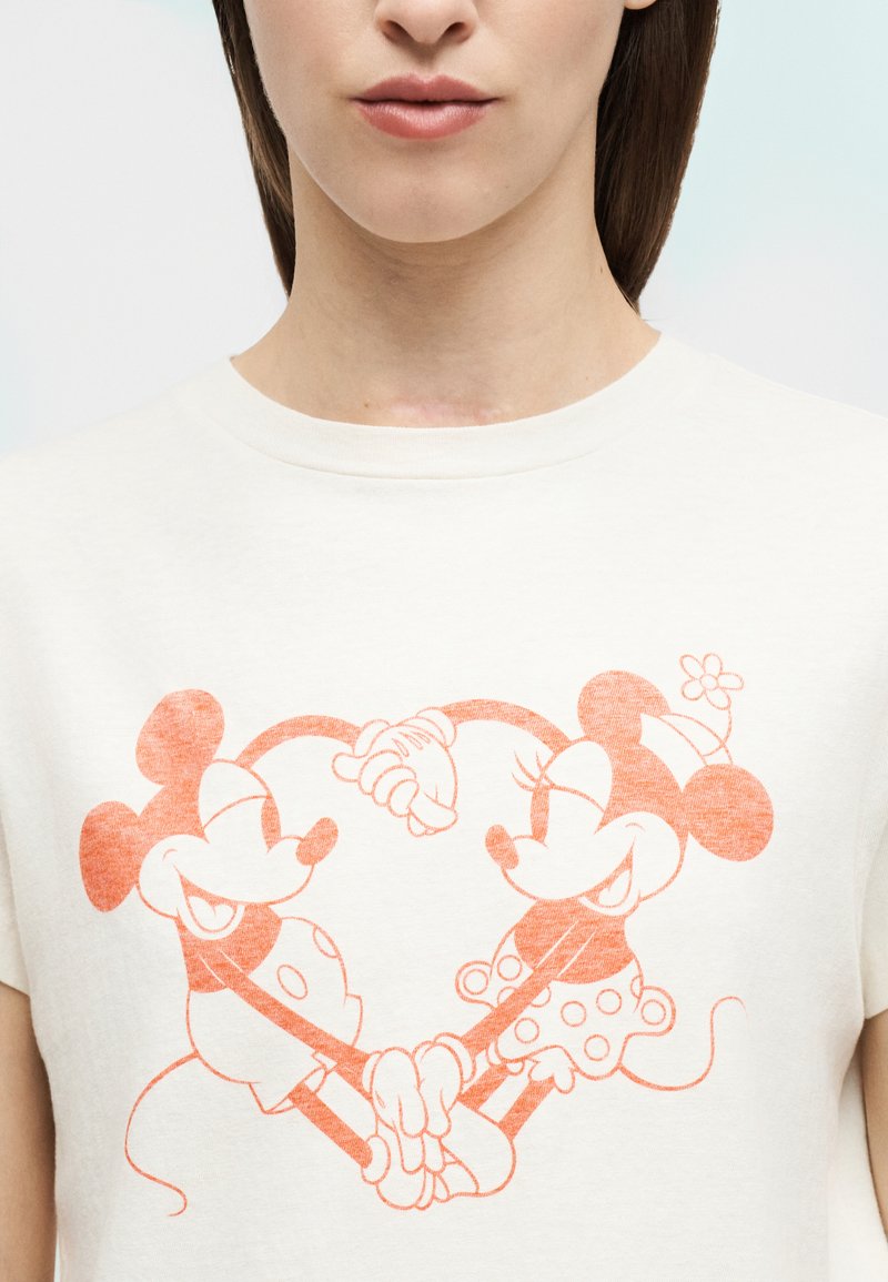 Frau trägt ein weißes T-Shirt mit orangefarbener Linienzeichnung von Mickey und Minnie Mouse, die Händchen halten und dabei eine Herzform zwischen ihnen bilden.
