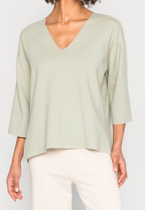 Femme portant un pull vert clair à col en V avec des manches trois-quarts et un pantalon blanc sur un fond uni.