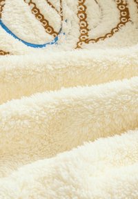 Krēmkrāsas mīksts segas ar maigu un pūkainu tekstūru; visā virsmā ir viļņaina brūnu un zilu toņu raksta izkārtojums.